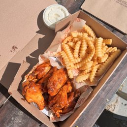 8 Wing Meal - Honey Hotter Flavor - A cardboard takeout container holds crispy chicken wings coated in sauce and crinkle-cut fries, with a small cup of dipping sauce nearby on a wooden surface.