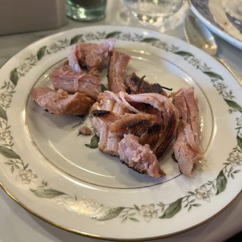 Toad in the Hole - Ham (pulled) - A decorative porcelain plate with a floral and gold trim border contains several pieces of cooked ham, some with charred edges. The plate is part of a table setting that includes a glass of water, a small jar, and other dishes in the background, indicating a meal setting. - Breakfast & Brunch Near Me - Eagle River, Wisconsin