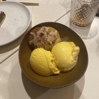 Joon - A small, round bowl holds two scoops of yellow ice cream, likely a flavor such as lemon or vanilla. Next to the ice cream is a cookie decorated with white icing and sprinkled with chopped pistachios. The bowl is on a white table, with a glass of beverage and a white plate nearby, suggesting a dessert setting. - Persian/Iranian Near Me - Vienna, Virginia