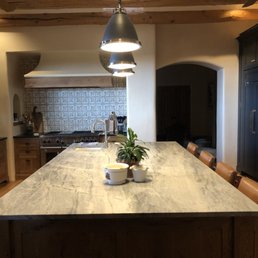 Strahle installed countertops and backsplash in kitchen remodel. - A kitchen features a large marble island with a plant and small bowls, black cabinets, and a tiled backsplash behind a stove.