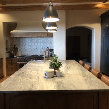 Strahle Tile & Granite - Strahle installed countertops and backsplash in kitchen remodel. - A kitchen features a large marble island with a plant and small bowls, black cabinets, and a tiled backsplash behind a stove. - Flooring Near Me - Albuquerque, New Mexico