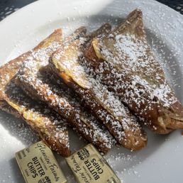 French Toast - A plate of French toast slices dusted with powdered sugar is displayed on a white plate. The slices are arranged overlapping each other, with butter packets placed at the edge of the plate. The setting appears to be outdoors, with sunlight illuminating the food and a textured surface visible in the background.
