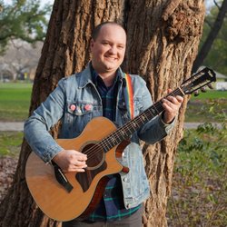 Eli Conley - Image Description: Eli, a white man, smiling and playing acoustic guitar in front of a tree. His guitar has a rainbow strap.