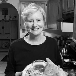 Happy Healthy Stellar 2021 ! - A woman with short, light-colored hair is smiling and holding a plate of food, which includes a large piece of bread, a bowl of sauce, and some salad. She is standing in a kitchen with wooden cabinets, a stove, and various kitchen utensils visible in the background. The image is in black and white.