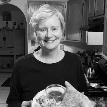 Davidos - Happy Healthy Stellar 2021 ! - A woman with short, light-colored hair is smiling and holding a plate of food, which includes a large piece of bread, a bowl of sauce, and some salad. She is standing in a kitchen with wooden cabinets, a stove, and various kitchen utensils visible in the background. The image is in black and white. - Pizza Near Me - Rio Rancho, New Mexico
