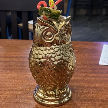 Fermentorium - A metallic gold owl-shaped glass contains a beverage, garnished with a slice of cucumber, a cherry, and colorful straws, placed on a wooden table. - Breweries Near Me - Cincinnati, Ohio
