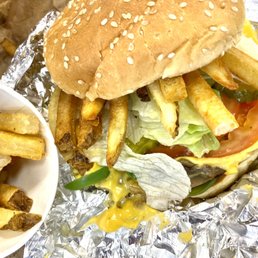 Veggie sandwich with cheese, added pickles and topped with some fries. - A veggie burger with a sesame seed bun contains lettuce, tomato, cheese, and pickles, topped with French fries. The burger is placed on crumpled aluminum foil, with additional fries served in a small bowl nearby. The ingredients include fresh vegetables and melted cheese, with the fries appearing crispy and golden.