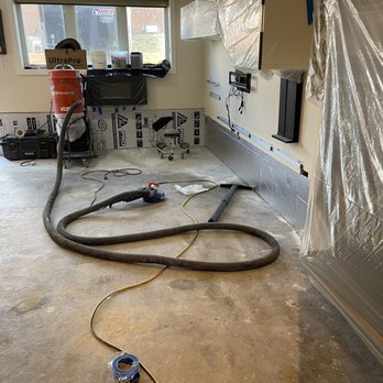 AD Technology - Before photo, floor grinding - The image depicts an indoor space under renovation, with construction equipment including vacuum hoses, a power tool, and electrical cords on the floor. The room has a window with natural light, and walls partially covered with plastic sheeting and insulation material. There are various tools and supplies, such as a toolbox, a stool, and construction materials, indicating ongoing work on the flooring or wall installation. - Flooring Near Me