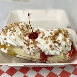 Banana Split - A banana split dessert features a sliced banana in a plastic takeout container, topped with generous whipped cream, chopped nuts, and a maraschino cherry. The container is placed on a red and white checkered paper, with a blurred background. The dessert includes fruit toppings such as strawberries and pineapple, along with chocolate syrup.