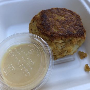 Harmony Bakery - Mock Crab Cake - A vegan crab cake with a golden-brown, crispy exterior is shown in a white takeout container. Next to it is a small, clear plastic cup filled with a light-colored dipping sauce. The container appears to be made of foam or similar material, and the crab cake is positioned on the right side of the image. - Gluten-Free Near Me - Baltimore, Maryland
