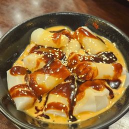 A black bowl contains rice rolls drizzled with caramel sauce and sprinkled with sesame seeds, served on a wooden table.