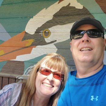 Turtle Bay Exploration Park - Outside the kids' play area - A man and woman are taking a selfie outdoors in front of a colorful mural of a bird painted on a wooden wall. The man is wearing a blue athletic shirt, a black cap, and sunglasses, while the woman has long hair, glasses, and is smiling. The mural depicts a bird with detailed features and bright colors, adding artistic context to the photo. - Museums Near Me - Redding, California