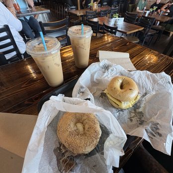 Yorgo's Bageldashery - Breakfast Short rib sandwich  Toasted Marshmallow and Rose Latte with Oat Milk BYOS  Brown Sugar Toasted Marshmallow Latte with Almond milk - Two iced coffee drinks and two bagels on parchment paper are placed on a wooden table in a busy restaurant with customers in the background. - Bagels Near Me - Norfolk, Virginia