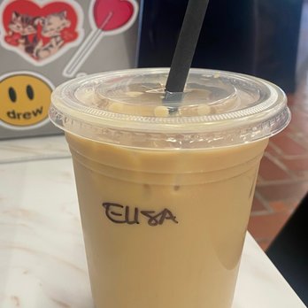 Edmonds Cafe - a cup of iced coffee - A clear plastic cup contains iced coffee with a light brown color, topped with a transparent lid and a black straw. The cup has the word 'ELISA' written on it. It is positioned on a white marble table, with a blurred background that includes a sticker or sign with cartoon characters and text. - Cafes Near Me - Berkeley, California
