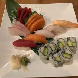 Sashimi - A square white plate holds a variety of sushi and sashimi, including slices of tuna, salmon, and white fish, along with shrimp nigiri. There are also avocado and crab rolls with rice and seaweed. Wasabi and pickled ginger are placed on the side. The presentation includes a green leaf and shredded daikon for garnish, set on a wooden table.