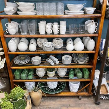 Bench492 - The image shows a wooden shelving unit with five shelves, filled with ceramic and glassware items. The top shelf has white bowls and glasses, while the second shelf contains white mugs, cups, and clear glasses. The middle shelves display decorative white vases, plates, and bowls, with some green ceramic vases and planters on the lower shelves. The setting appears to be a retail store or boutique specializing in home decor and tableware. - Home Decor Near Me - Glen Ellyn, Illinois