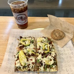 Italian Stallion Pizza - A rectangular vegetable pizza topped with green peppers, onions, and melted cheese is placed on branded paper on a wooden table. To the right, there is a chocolate chip cookie on a piece of parchment paper. Behind the pizza, a clear plastic cup filled with iced tea and ice cubes, bearing the Azzip Pizza logo, is visible. The background shows a reflective surface, possibly a window or glass partition.