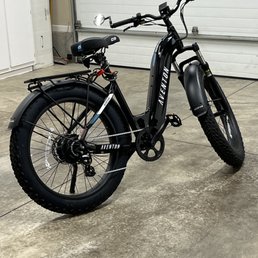 A black Aventon electric bike with fat tires is positioned in a garage, featuring a rear rack, disc brakes, and a display on the handlebar.