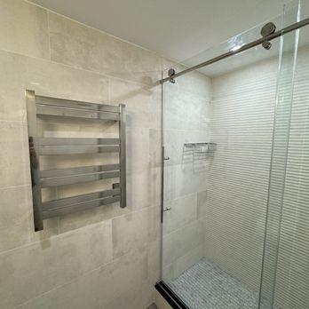 Porcelanosa - A shower space featuring a glass door, metal towel rack, and tiled walls with a textured pattern, in a contemporary bathroom. - Kitchen & Bath Near Me - Paramus, New Jersey