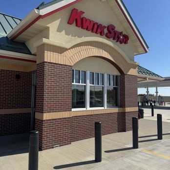 Kwik Star - The image shows the exterior of a Kwik Star convenience store with a brick facade and a large window. The building has a gabled roof with white trim and a red and white sign displaying the Kwik Star logo. The surrounding area includes a paved sidewalk with black bollards and parking spaces, and a clear blue sky overhead. - Gas Stations Near Me - Mitchellville, Iowa