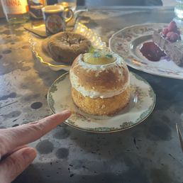 A dessert with a meringue topping, garnished with lemon and small green leaves, sits on a decorative floral-patterned plate. The table has other desserts, drinks, and condiments visible in the background, indicating a dining setting.