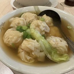 Shrimp wonton - A bowl of shrimp wonton soup features several dumplings filled with shrimp, garnished with chopped green onions and a leafy vegetable, all immersed in a clear broth. The bowl is placed on a wooden table, with a black spoon resting inside. In the background, there is another bowl and a small jar, indicating a dining setting at a restaurant.