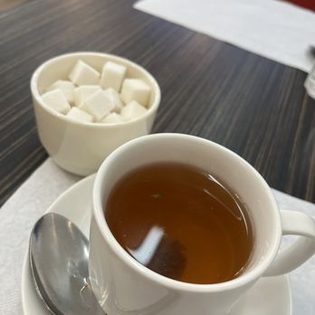 Kabul Corner - Tea - A white ceramic cup filled with tea is placed on a matching saucer, with a metal spoon resting on the saucer's edge. Behind the cup, there is a small bowl filled with sugar cubes. The setting is on a dark wooden table, with a white paper napkin and a red chair visible in the background. The scene suggests a casual dining environment at Kabul Corner in Tucson, AZ, offering traditional Afghan tea and sweets. - Afghan Near Me - Tucson, Arizona