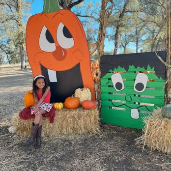 Fowler Ranch - A girl in a red dress and cowboy boots sits on hay bales beside fall decorations, including pumpkins and a large pumpkin cutout with a face, and a painted wooden Frankenstein figure. - Pumpkin Patches Near Me - Lincoln, California