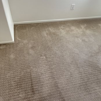 Stanley Steemer - The image depicts a beige carpet with visible dirt, stains, and wear marks in a room corner. The carpet is adjacent to a white wall with a power outlet and a white baseboard. The room appears to be in an indoor residential or office setting, with the carpet showing signs of use and needing cleaning or replacement. - Carpet Cleaning Near Me - Oxnard, California