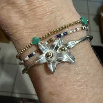 Eden - The image shows a close-up of a wrist wearing multiple bracelets. One bracelet is a silver band with a starfish-shaped charm, while others include beaded bands with small turquoise, gold, and multicolored beads. The background includes tiled flooring and part of a chair, indicating an indoor setting. - Jewelry Near Me - Dennis, Massachusetts