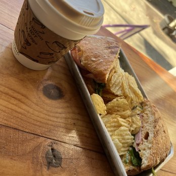 Victory Point Cafe - The king in the north - A meal consisting of a sandwich with visible ingredients such as greens and possibly meat, served alongside ridged potato chips in a metal tray. A disposable coffee cup with a cardboard sleeve and plastic lid is placed nearby on a wooden table. The setting appears to be a casual dining or cafe environment with natural lighting. - Coffee & Tea Near Me - Berkeley, California