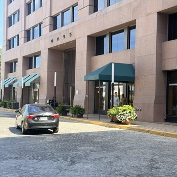 Joon - The image shows a modern, multi-story building with large, reflective windows and a beige stone facade. The entrance features a green awning with the word 'LOV' visible, and there are potted plants and benches near the entrance. A black car is parked on a cobblestone street in front of the building, with two people visible near the entrance. The scene appears to be in an urban area with clear weather. - Persian/Iranian Near Me - Vienna, Virginia