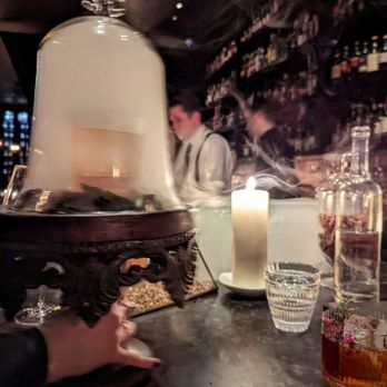 BarChef - Fantastic bar, fantastic bartenders, great service. - A hand holds a glass dome over a dessert on a bar counter, with a lit candle, a glass of water, and bottles on shelves behind the bar. - Lounges Near Me - Toronto, Ontario