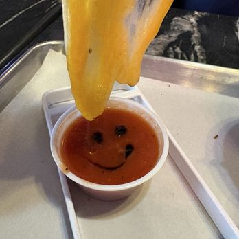 Grilled Cheese Gallery - Yummy Tomato Basil Soup (Dipper) - A grilled cheese sandwich is being dipped into a cup of tomato soup, which has a smiley face drawn on its surface, on a tray. - Ice Cream & Frozen Yogurt Near Me - St. Augustine, Florida