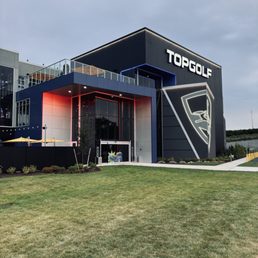 The image shows the exterior of a Topgolf facility with modern design, neon accents, and a large illuminated logo, set against a cloudy sky.