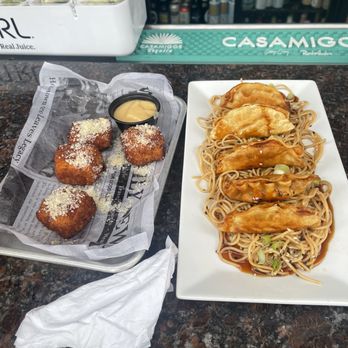 Craftu - The image shows a restaurant counter with two dishes: a tray of four fried croquettes topped with grated cheese and a small container of dipping sauce, and a rectangular plate of five chicken pieces served over noodles with sauce and chopped green onions. The background includes beverage refrigerators stocked with drinks and a green Casamigos tequila sign. The counter surface is dark and polished. - Bars Near Me - Lynbrook, New York