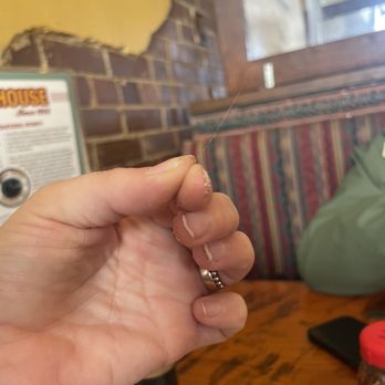 Pizza House - Hair in my pizza - A close-up of a person's hand, showing dirt under the fingernails, holding a thin, nearly invisible strand of spider silk. The background includes a brick wall, a wooden table, and restaurant decor, indicating the photo was taken inside a casual dining establishment. - Pizza Near Me - Springfield, Missouri