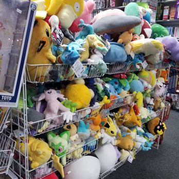Toy Vault - Stuffed pokemon and other critters - A collection of Pokémon plush toys is displayed on wire racks inside a retail store. The plush toys vary in size, color, and character, including Pikachu, Bulbasaur, and other Pokémon. The store shelves in the background contain boxed merchandise and electronic items, indicating a toy and collectibles retail environment. The plush toys are organized in multiple rows, with some larger plush toys placed at the top and smaller ones below. - Toy Stores Near Me - Warwick, Rhode Island