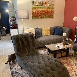 Living room. Chaise lounge was cleaned as well as the carpets. - The image shows a living room with a gray sofa adorned with yellow, navy, and dark gray pillows, and a tufted armchair in the foreground. A small coffee table holds a candle, a tray, and decorative items. There are cats visible, one near the armchair and another partially visible on the right. The room has a colorful area rug, floral arrangements, and a large wall art piece depicting a landscape. The background includes a dining area with a table, chairs, and additional decor, creating a warm, inviting atmosphere.