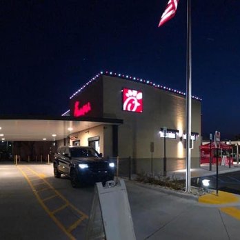 Chick-fil-A - Exterior - The image depicts the exterior of a Chick-fil-A restaurant at night, featuring illuminated signage with the Chick-fil-A logo. The building has a drive-thru lane with a vehicle present, and the area is lit by exterior lights. An American flag is visible on a flagpole near the entrance, and the sky is dark, indicating nighttime. The scene includes parking spaces and a sidewalk area. - Fast Food Near Me - Glendale, Wisconsin