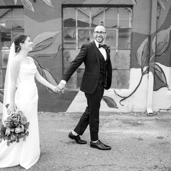 Bella Tailor - A bride and groom walk outdoors holding hands in front of a mural wall featuring painted leaves and abstract designs. The bride wears a white wedding gown and veil, holding a bouquet of flowers, while the groom is dressed in a black tuxedo with a bow tie and glasses, smiling as he looks ahead. The ground is paved, and the mural provides a colorful background for the scene. - Sewing & Alterations Near Me - Denver, Colorado