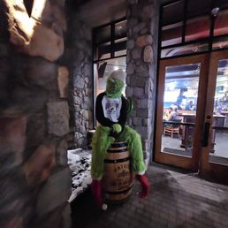 Entrance - A person in a Grinch costume, with green fur and a mask, is seated on a barrel outside a building with stone walls and glass doors.