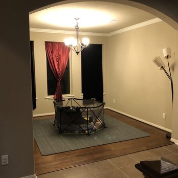 Plus58 Remodeling - Downstairs dining room before - A dining room features a chandelier, two windows with curtains, and a floor lamp, with a playpen on a rug in the center. - General Contractors Near Me