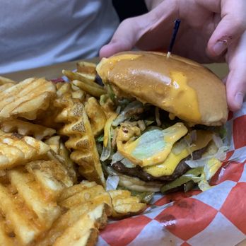AJ's - Boom Boom Burger - A cheeseburger features a bun, melted cheese, lettuce, and sauce, with a hand holding it. The burger is served with a side of waffle fries on a red and white checkered paper-lined tray, in a casual dining setting. - American Near Me - West Lafayette, Indiana