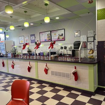 Yogurt Time - Toppings bar... Rush disappeared...worker really on top of cleanliness!   Gentleman working is thorough! - The image shows the interior of a frozen yogurt shop with a long counter decorated for the holiday season, including Christmas stockings hanging along the front. The ceiling has green pendant lights, and the walls are decorated with framed menus and holiday-themed accents. The floor has a black and white checkered pattern, and there are seating areas with red chairs visible in the foreground. The shop appears clean and festive, with a focus on holiday decor. - Ice Cream & Frozen Yogurt Near Me - Commack, New York