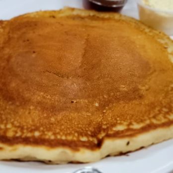 Cormier’s Kitchen - Pancake - A thick pancake with a golden-brown surface is presented on a white plate, accompanied by small containers of syrup, butter, and other toppings. - Southern Near Me - Spring, Texas