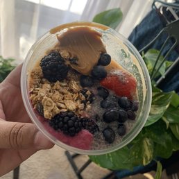 A hand holds a clear plastic bowl filled with a smoothie topped with granola, blackberries, blueberries, and a strawberry. The background shows a window with curtains, some indoor plants, and a clothing rack. The setting appears to be indoors near a window with natural light.
