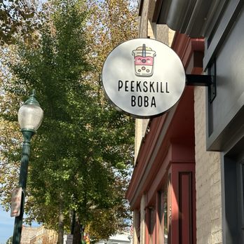 Peekskill Boba - A round sign with a bubble tea illustration and the text 'Peekskill Boba' is mounted on the exterior of a building, with trees and a street scene in the background. - Bubble Tea Near Me - Peekskill, New York