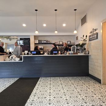 The Southern Cafe - The photo depicts the interior of a cafe with a dark wood counter where staff are preparing orders. Behind the counter, shelves hold jars and bottles, and a chalkboard menu is visible. Customers are seated in the background, and the floor features patterned tiles. Pendant lights hang from the ceiling, illuminating the space. To the right, a doorway leads to another area, with a sign indicating restrooms. - Southern Near Me - Roselle, Illinois
