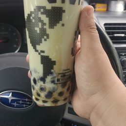 Honeydew milk tea with tapioca boba! Their boba is the best! - A person is holding a bubble tea with tapioca pearls at the bottom, in a clear plastic cup with a pixelated world map design and partial text. The background shows the interior of a vehicle, including a steering wheel with a Subaru logo and part of the dashboard. The drink appears to be a milk tea or similar beverage, with a light-colored liquid and dark tapioca pearls.
